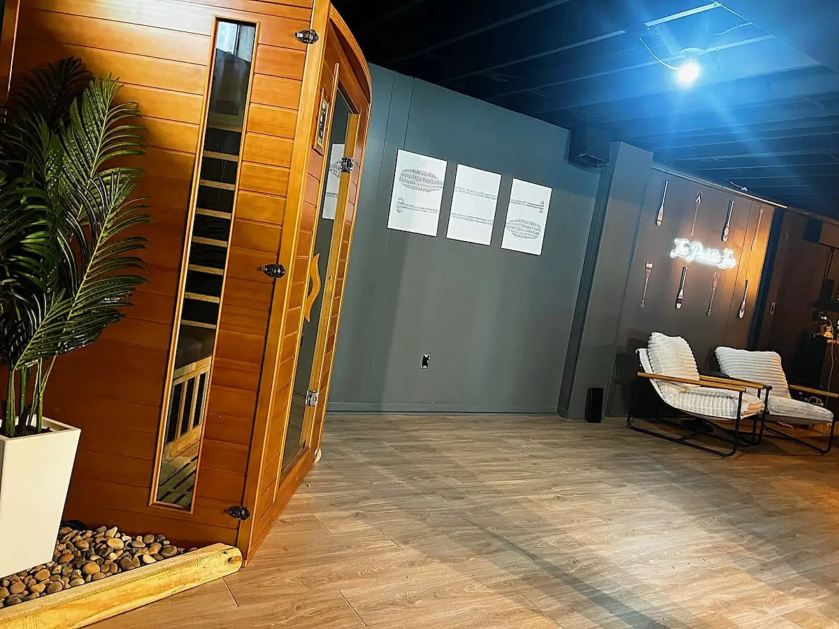 A wood-panelled sauna cabin in the basement wellness area, beside two loungers under a neon sign reading The Paddle Inn