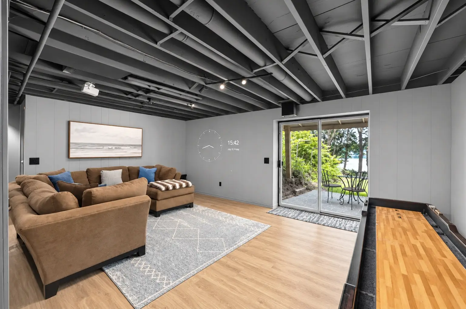 Basement lounge with a sectional sofa, a ceiling-mounted projector, a shuffleboard table along the right wall, and sliding doors out to a patio