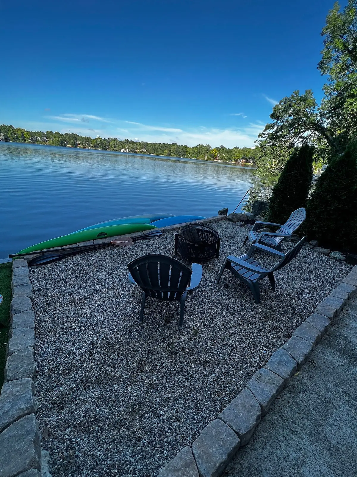 Gravel fire pit area with Adirondack chairs at the water's edge, kayaks pulled up alongside