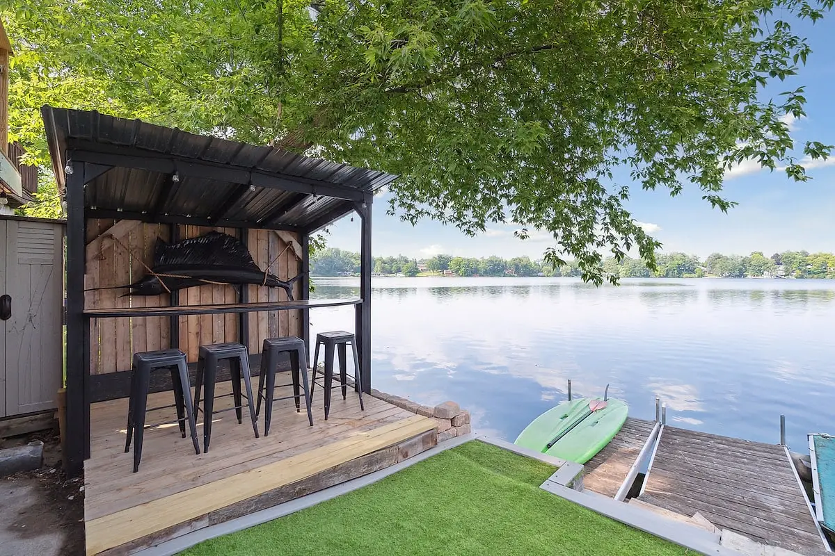 The Black Marlin Bar, an open-sided tiki bar with a mounted black marlin and metal stools, beside paddleboards on the dock and the lake beyond